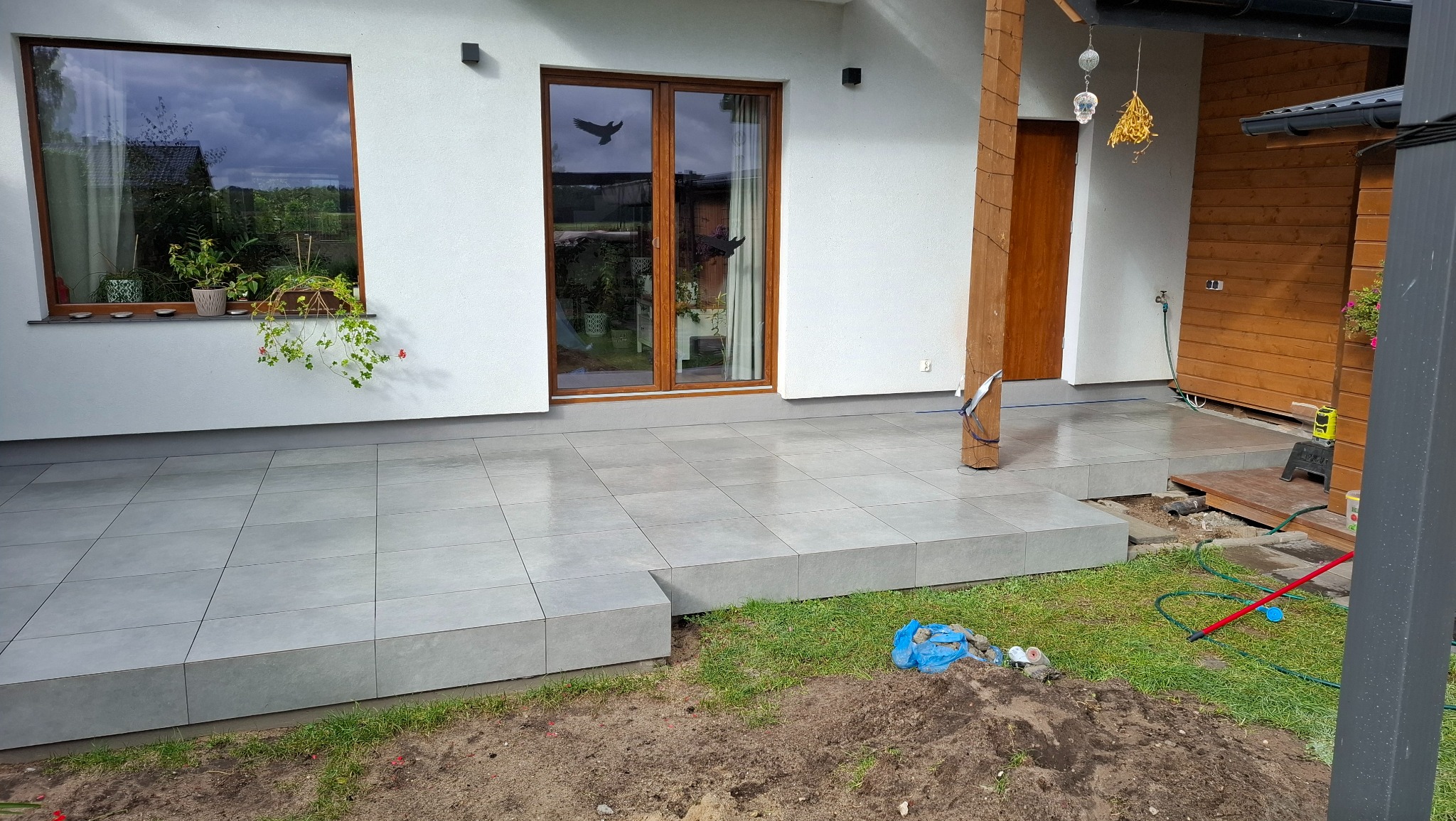 Wykończony taras z szarych, kwadratowych płytek, z widocznym fragmentem ogrodu i elewacją domu z oknami. Część płytek jeszcze nieułożona.