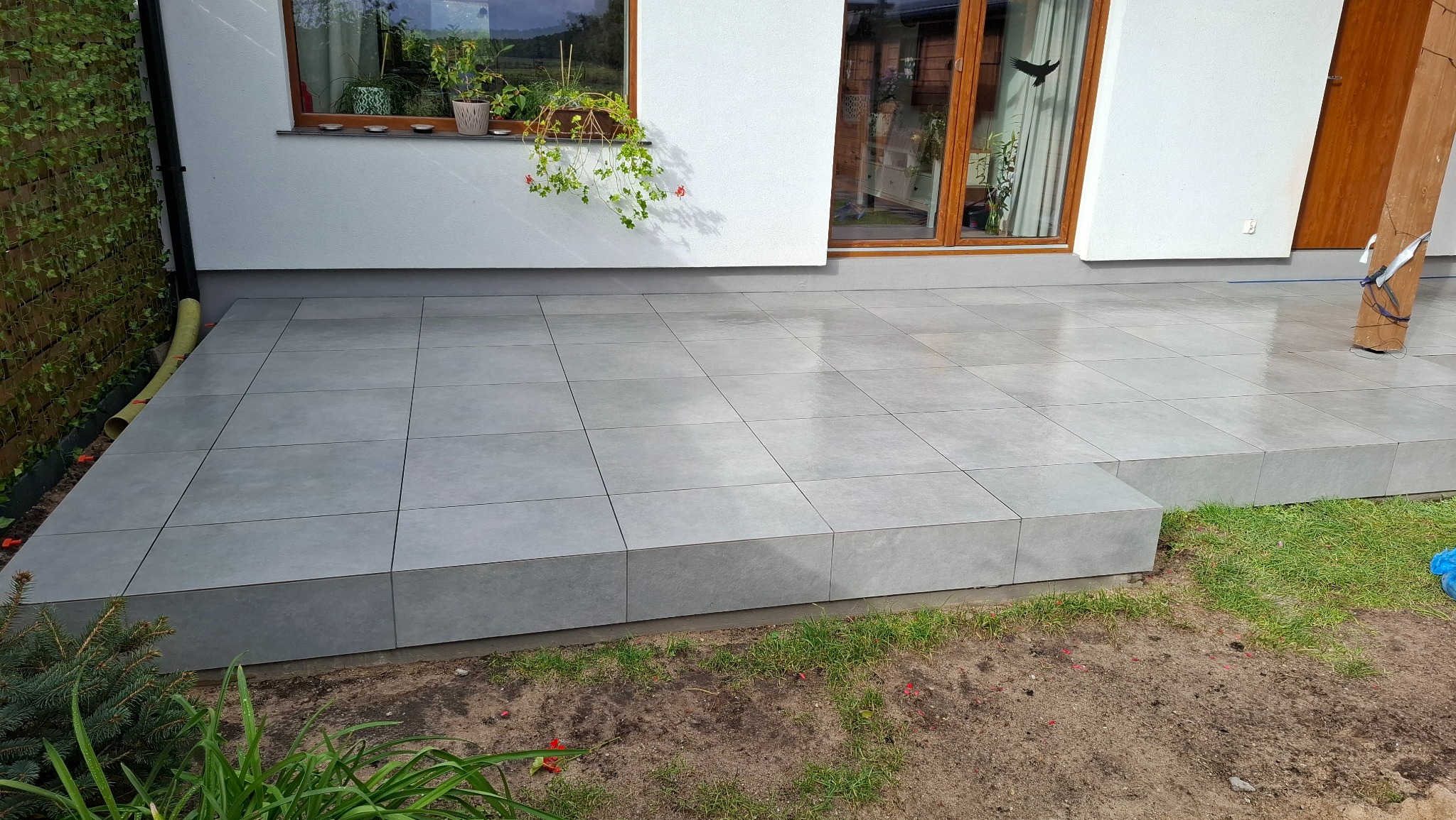 Wyłożony szarymi płytkami taras z niskim stopniem, widok z dołu, zieleń w tle, okno z kwiatami na ścianie budynku. Minimalistyczny design.