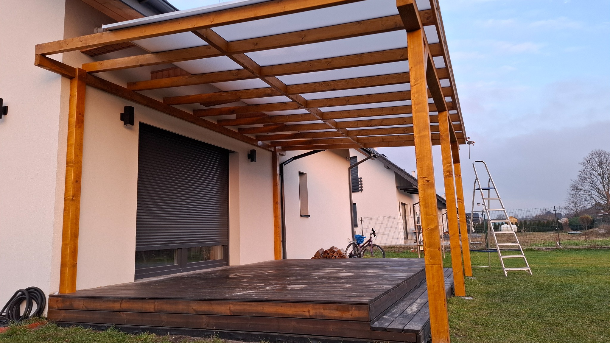 Drewniana pergola z zadaszeniem z poliwęglanu przy białym domu z szarą roletą. Widoczna drabina i rower w tle. Całość w naturalnej kolorystyce.