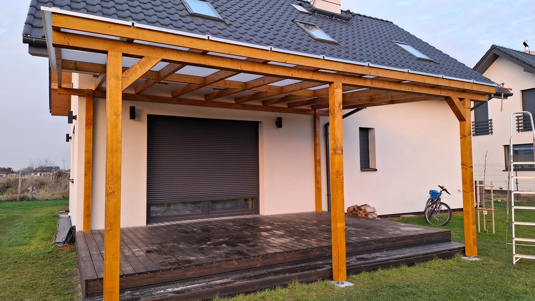 Drewniana pergola z zadaszeniem przy nowoczesnym domu z szarą roletą i rowerem. Widoczne schody z desek i trawnik. Ujęcie z poziomu gruntu, w tle sąsiedni budynek.