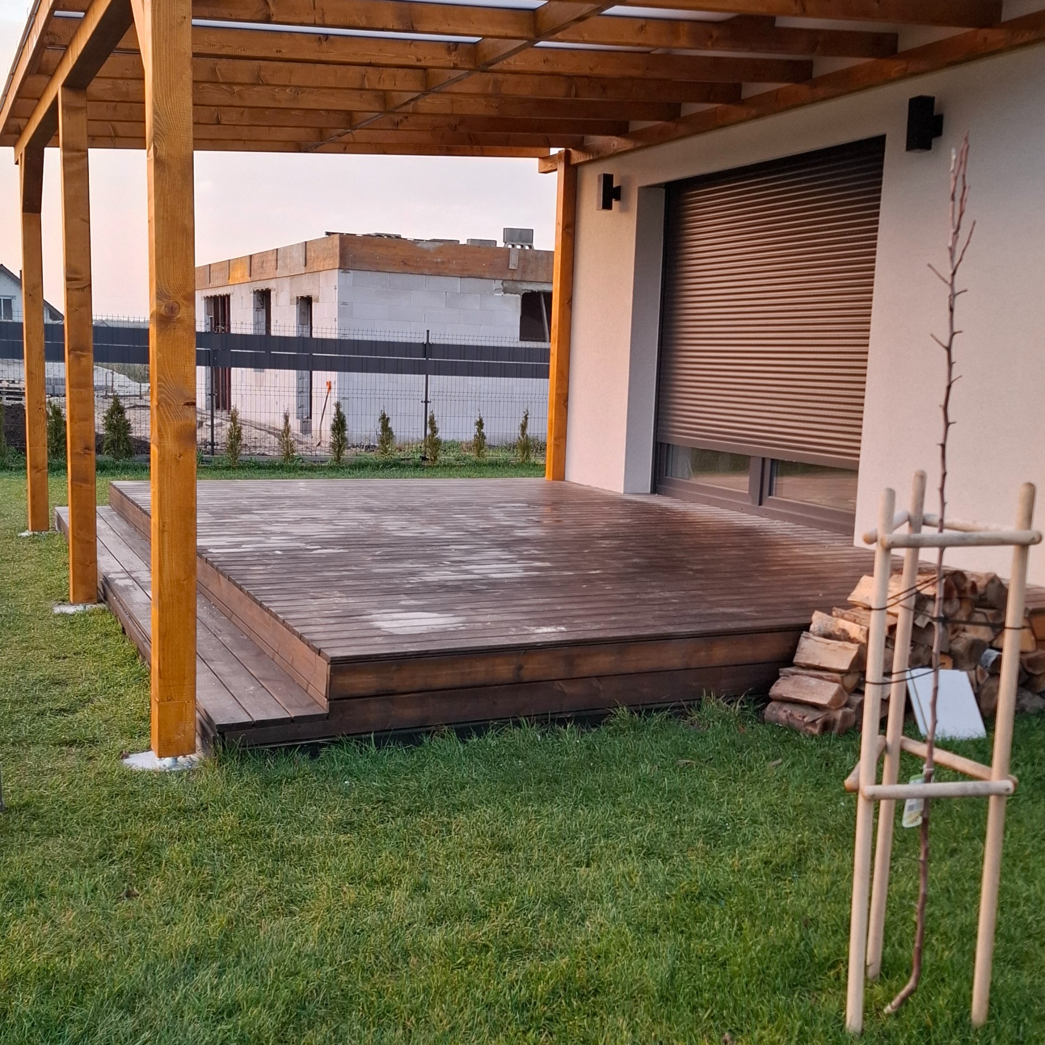 Drewniany taras z zadaszeniem przy domu z roletami zewnętrznymi. W tle budynek w trakcie budowy i zielony trawnik. Widok na zewnątrz.