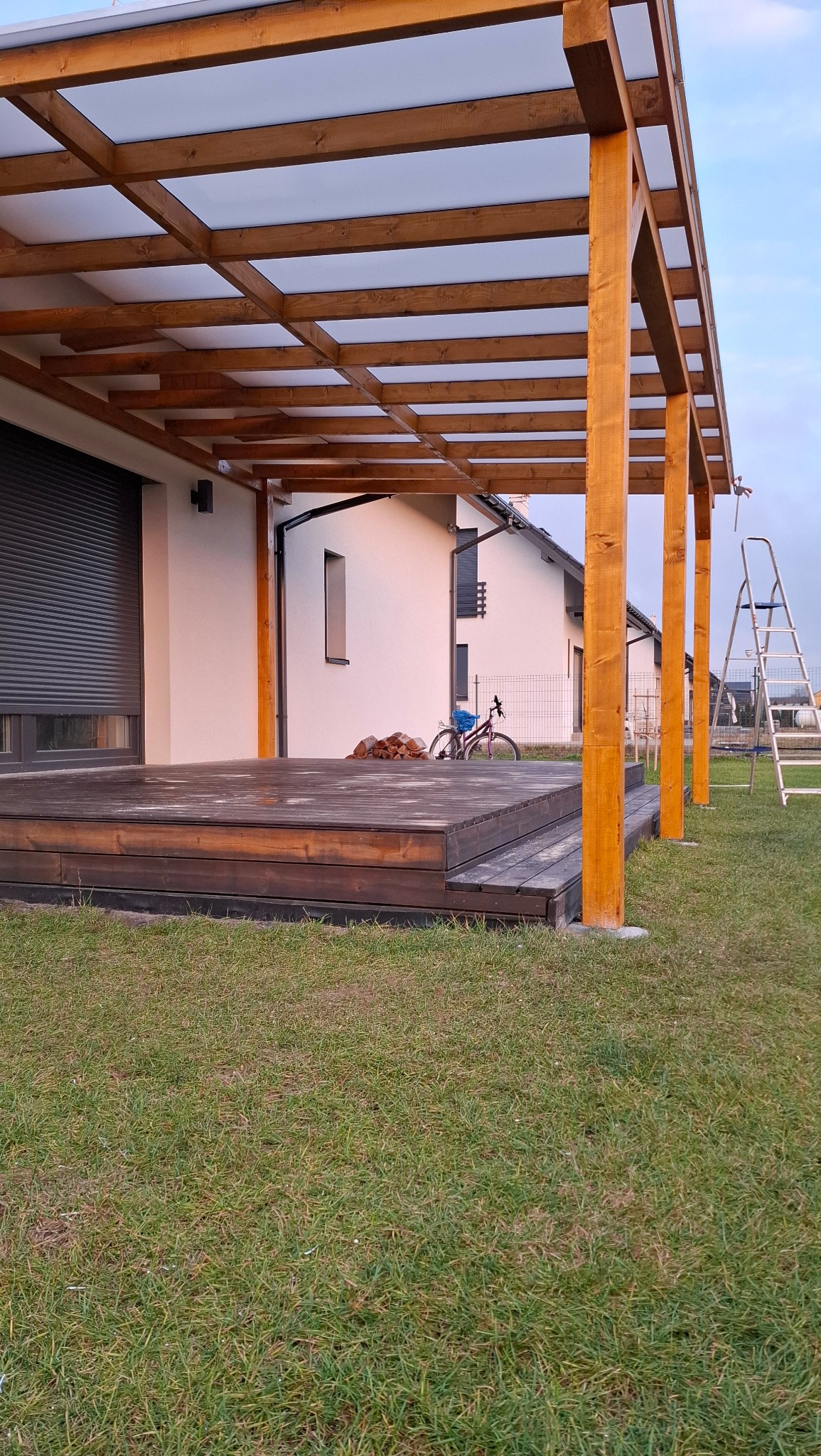 Drewniany taras z zadaszeniem z poliwęglanu przy białym domu. Na trawniku rower i drabina. Widok na sąsiedni dom. Całość w spokojnej, naturalnej kolorystyce.