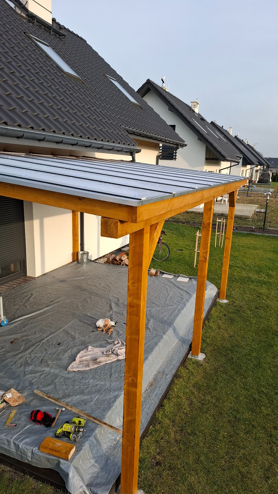 Drewniana konstrukcja z zadaszeniem na posesji, widoczne narzędzia i materiały budowlane, w tle domy jednorodzinne z ciemnymi dachami.