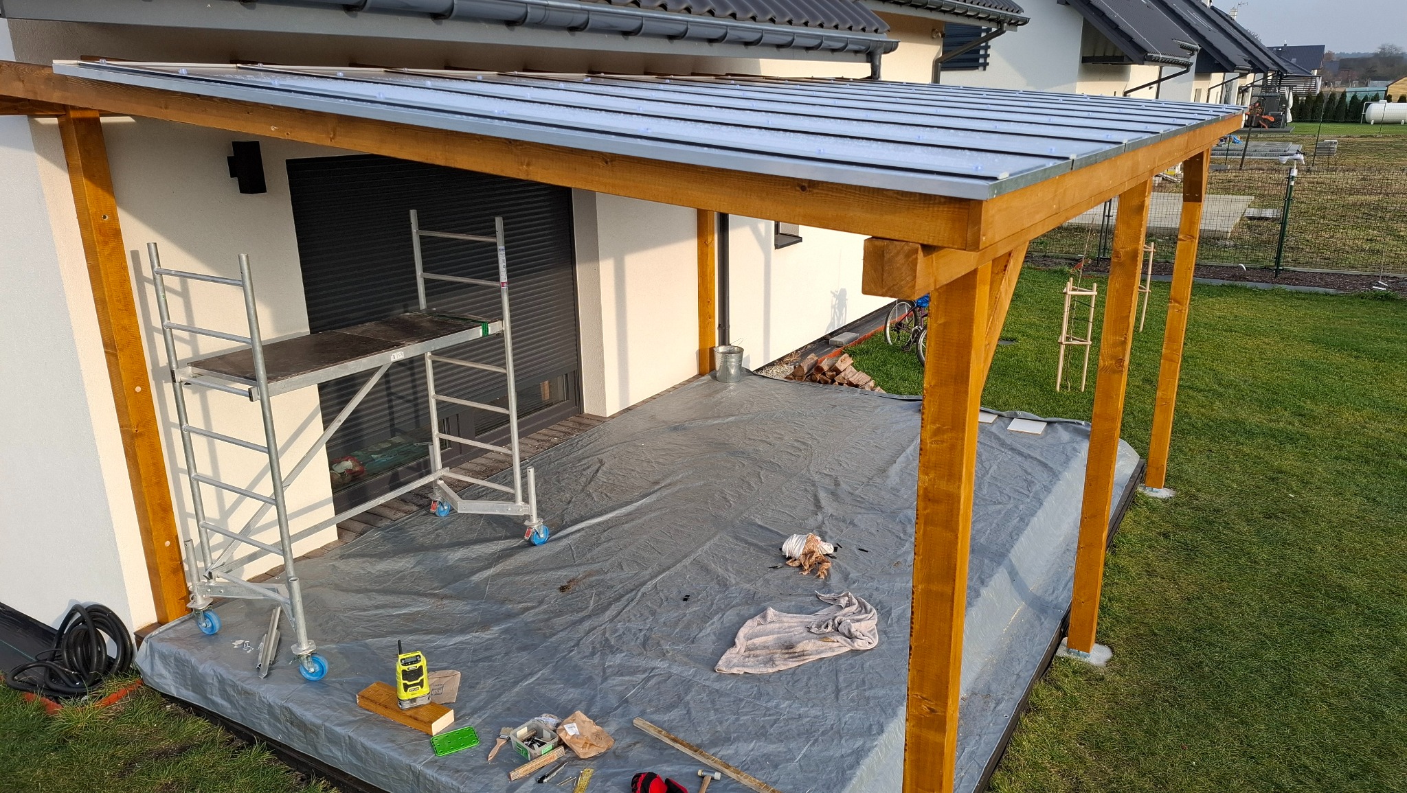Drewniana konstrukcja z metalowym dachem w trakcie budowy, widoczne rusztowanie i narzędzia na zabezpieczonej folią podłodze. Prace wykończeniowe w ogrodzie.