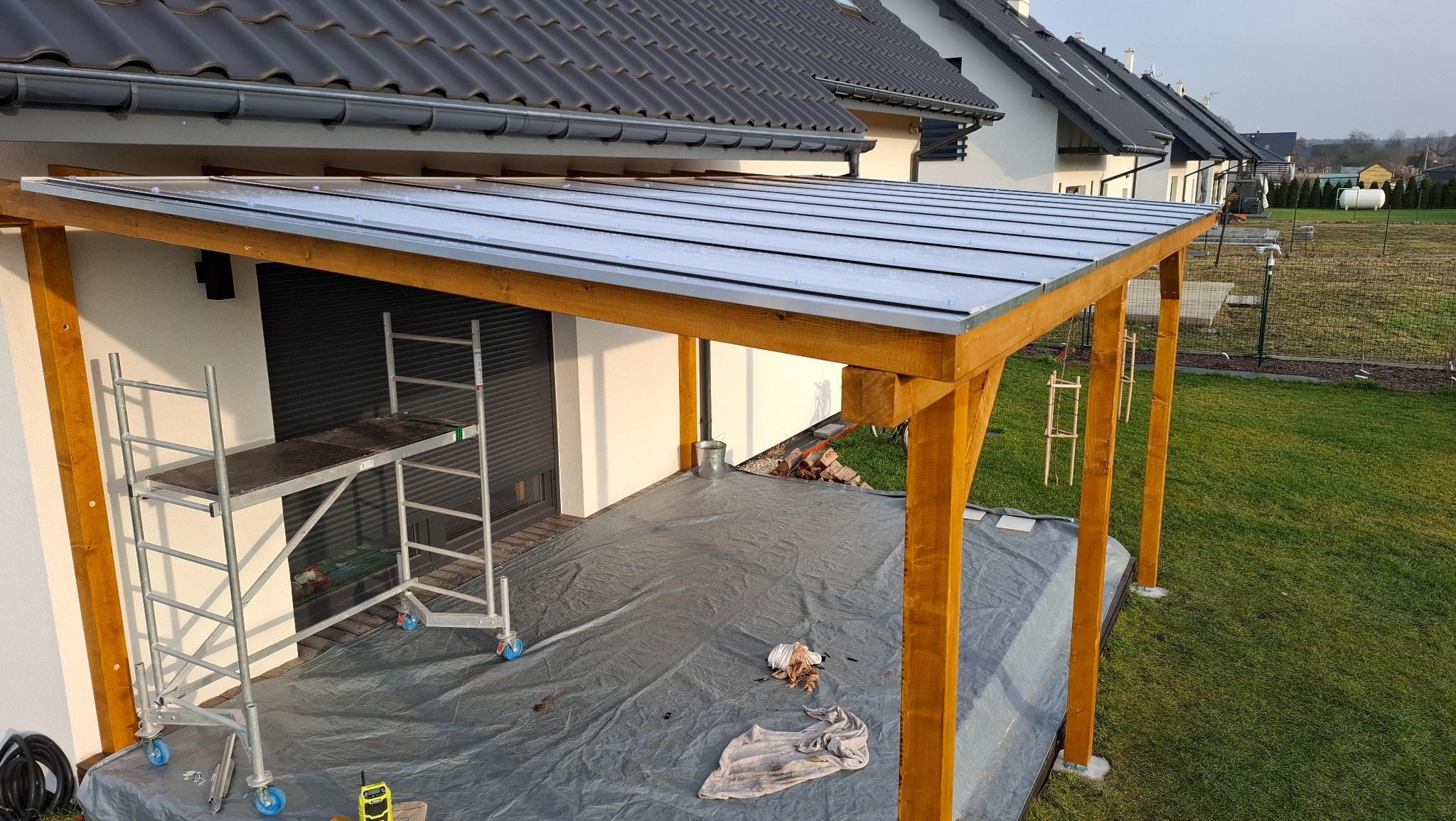 Drewniana konstrukcja z zadaszeniem na posesji, widok z góry. Na podłożu folia ochronna, obok rusztowanie. W tle domy jednorodzinne i trawnik.