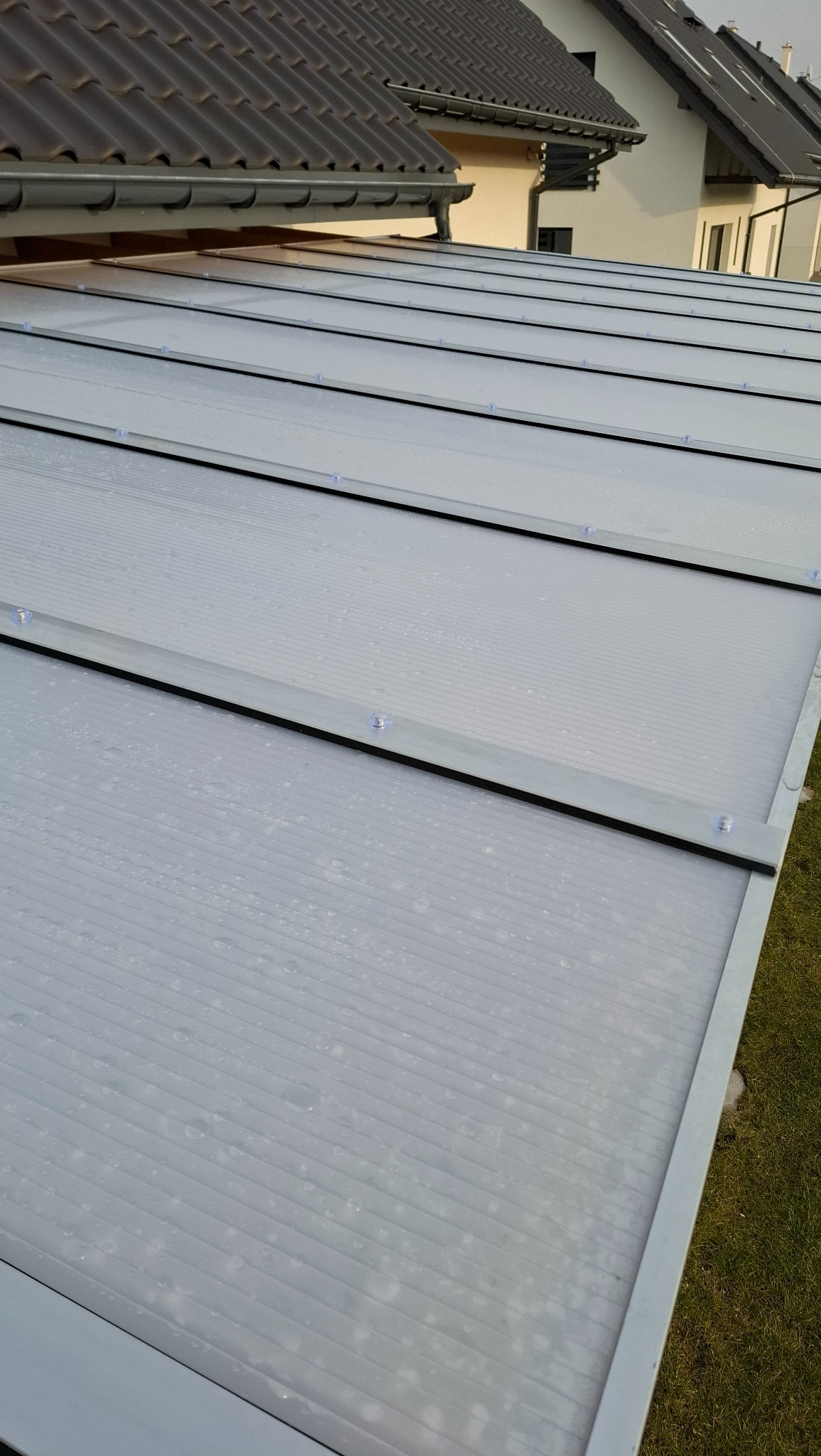 Przezroczyste panele dachowe z widocznymi kroplami wody, zamontowane na metalowej konstrukcji, z fragmentem dachu budynku w tle.