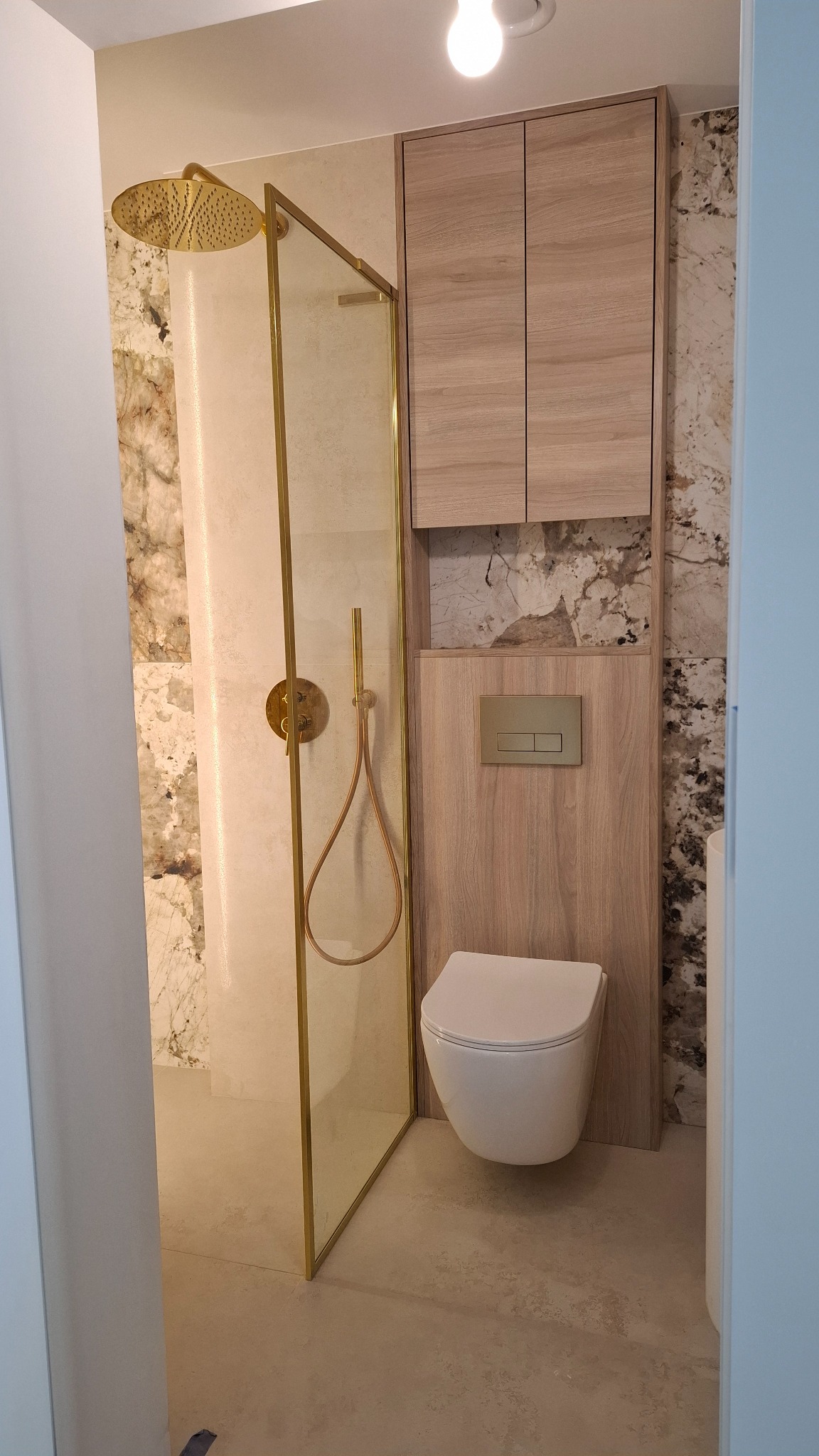 Nowoczesna łazienka po remoncie: kabina prysznicowa ze złotymi detalami, podwieszana toaleta i szafka w odcieniach drewna na tle marmurowych płytek.