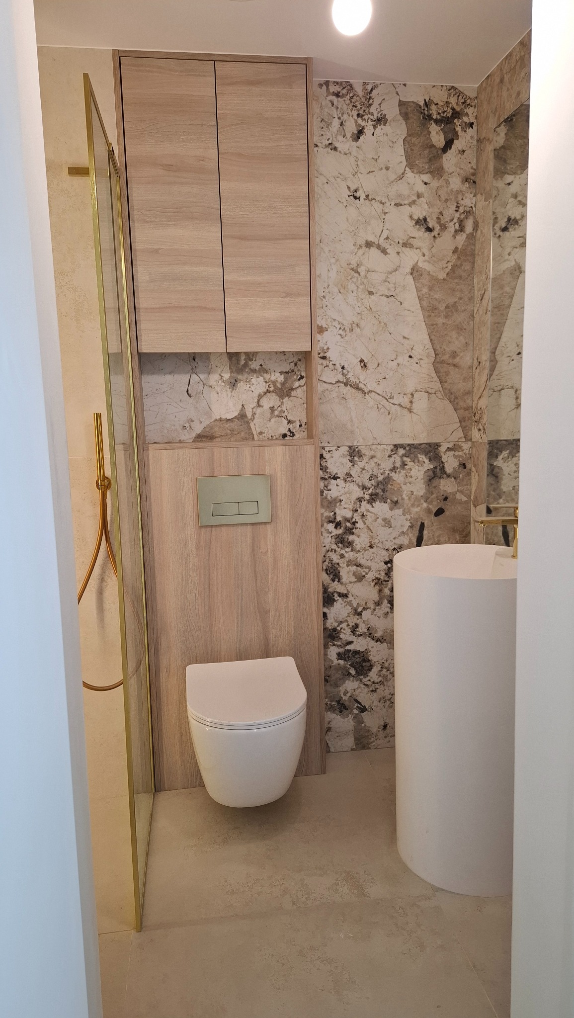 Nowoczesna łazienka z marmurową ścianą, wiszącą toaletą i kabiną prysznicową ze złotymi detalami. Minimalistyczny design, jasne kolory, eleganckie wykończenie.