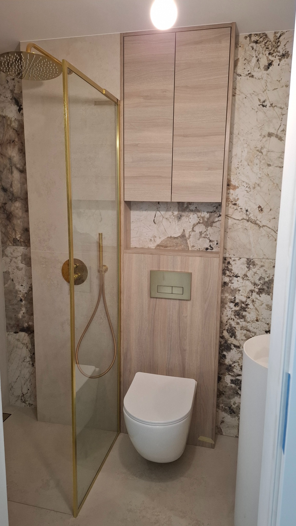 Nowoczesna łazienka po remoncie: kabina prysznicowa ze złotymi detalami, podwieszana toaleta i marmurowe płytki. Minimalistyczny design z elementami drewna.