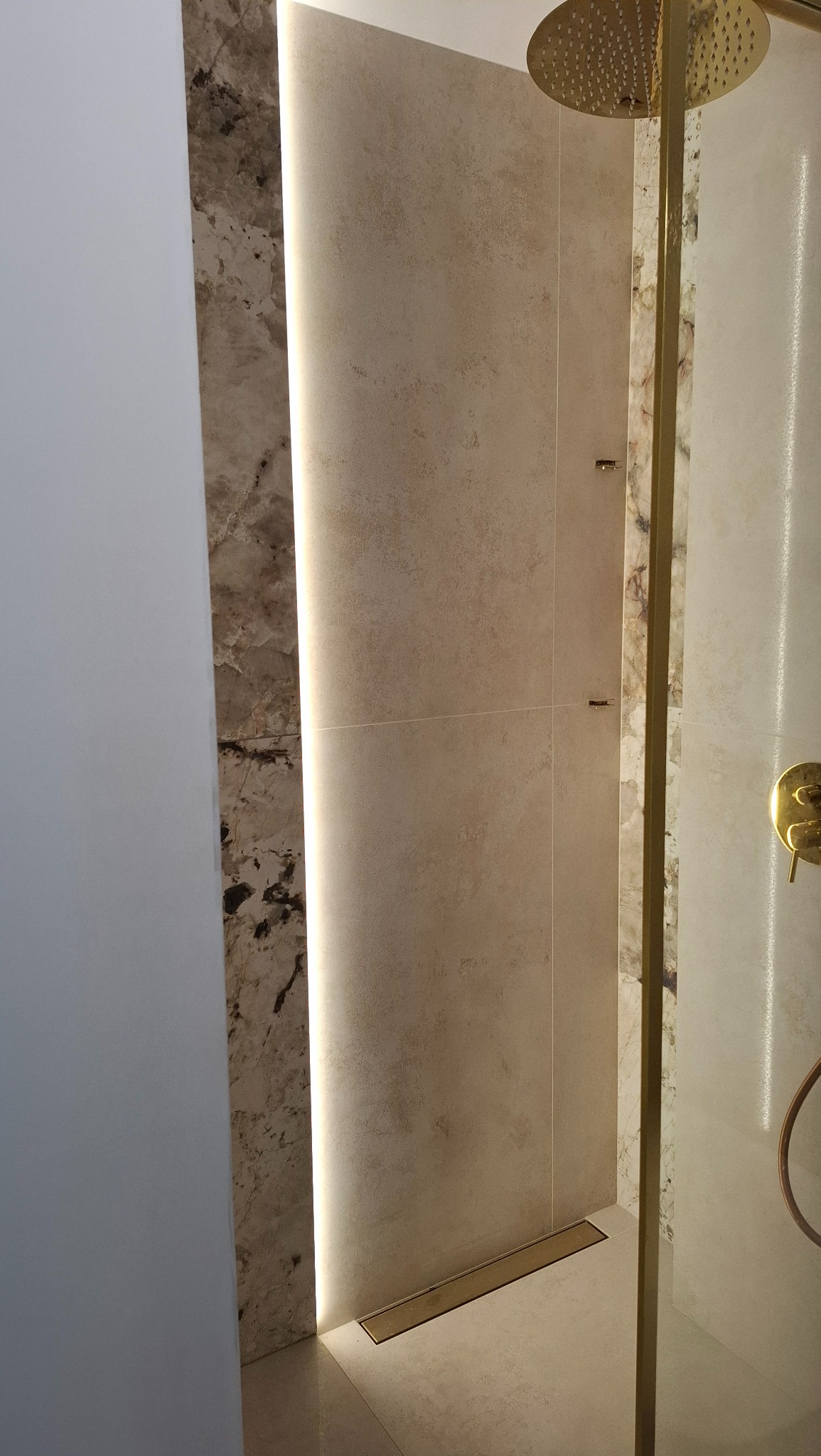 Beżowa kabina prysznicowa z odpływem liniowym i złotą armaturą. Podświetlane płytki imitujące marmur. Nowoczesny design.