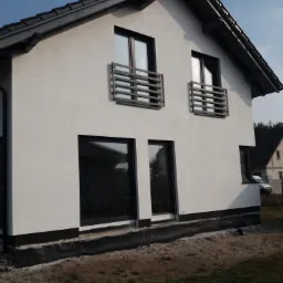 Boczny widok dwukondygnacyjnego domu z białymi ścianami, czarną dachówką i ciemnymi oknami. Widoczne balustrady balkonowe z poziomych belek i czarna folia fundamentowa wzdłuż podstawy ściany.