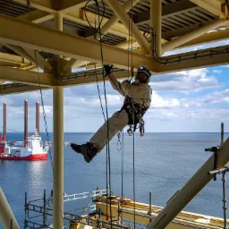 Pracownik w kombinezonie i kasku, zabezpieczony linami, wykonuje prace na wysokości na platformie wiertniczej, w tle widoczny statek i morze.