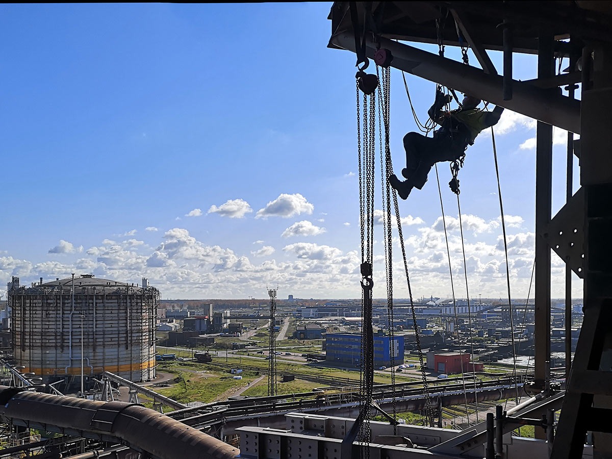 Pracownik zabezpieczony linami wykonuje zadania na wysokości na tle zbiornika przemysłowego i zabudowy miejskiej w słoneczny dzień.