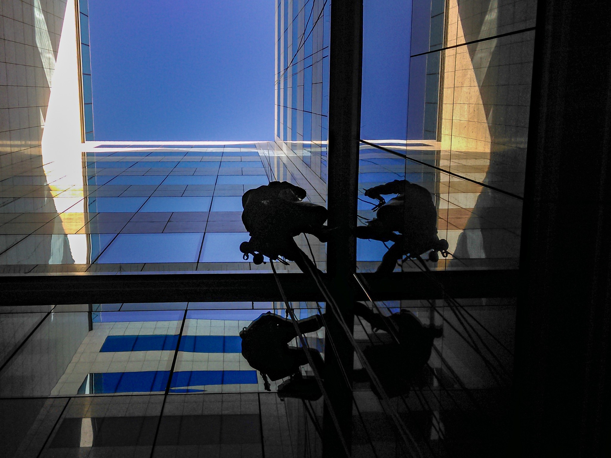 Silhouettes of two workers suspended by ropes against a backdrop of a modern glass office building and a bright blue sky, viewed from below.