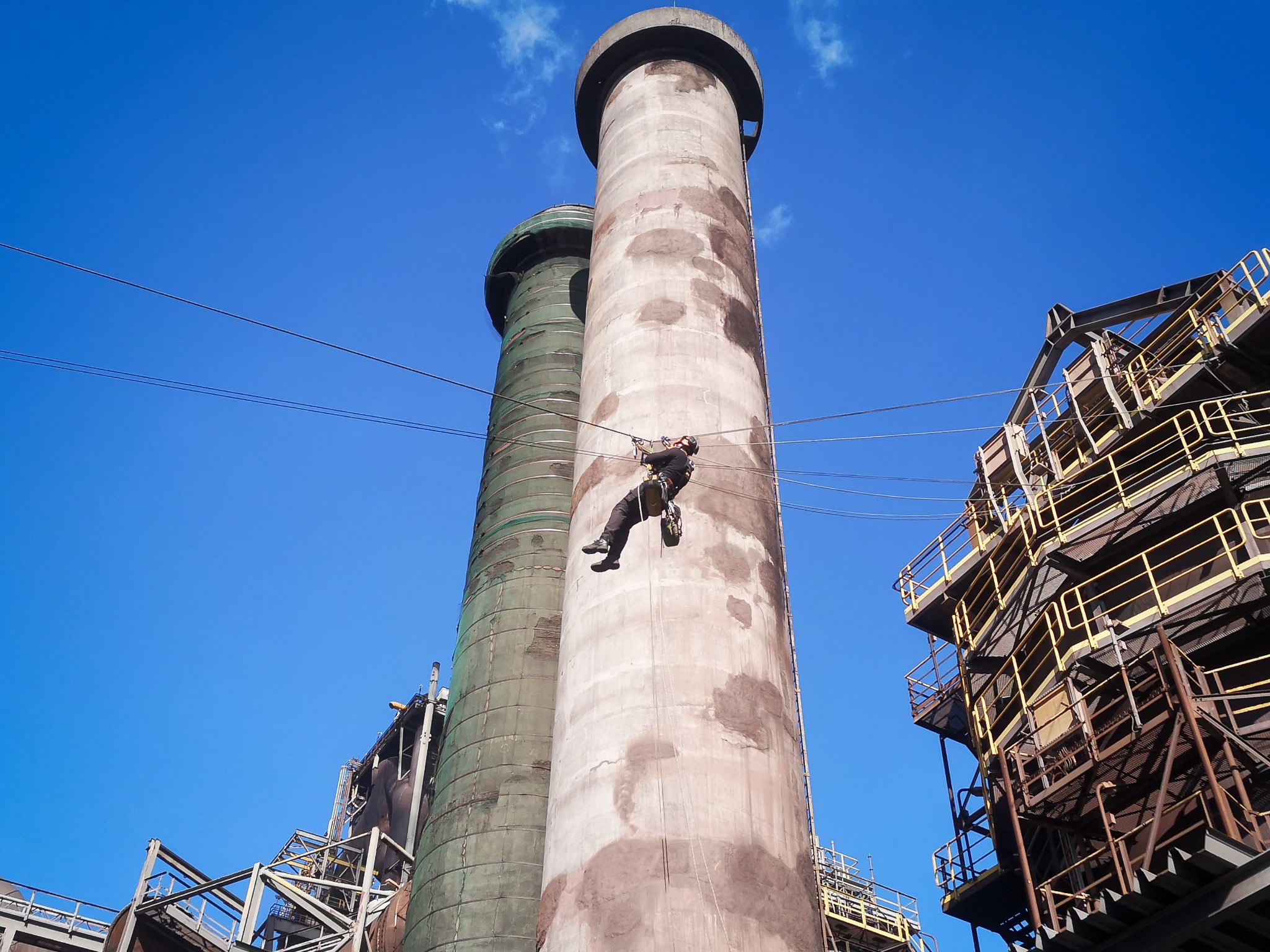 Pracownik wysokościowy zjeżdża na linie po betonowym kominie przemysłowym na tle błękitnego nieba, widoczne są metalowe konstrukcje i drugi komin.