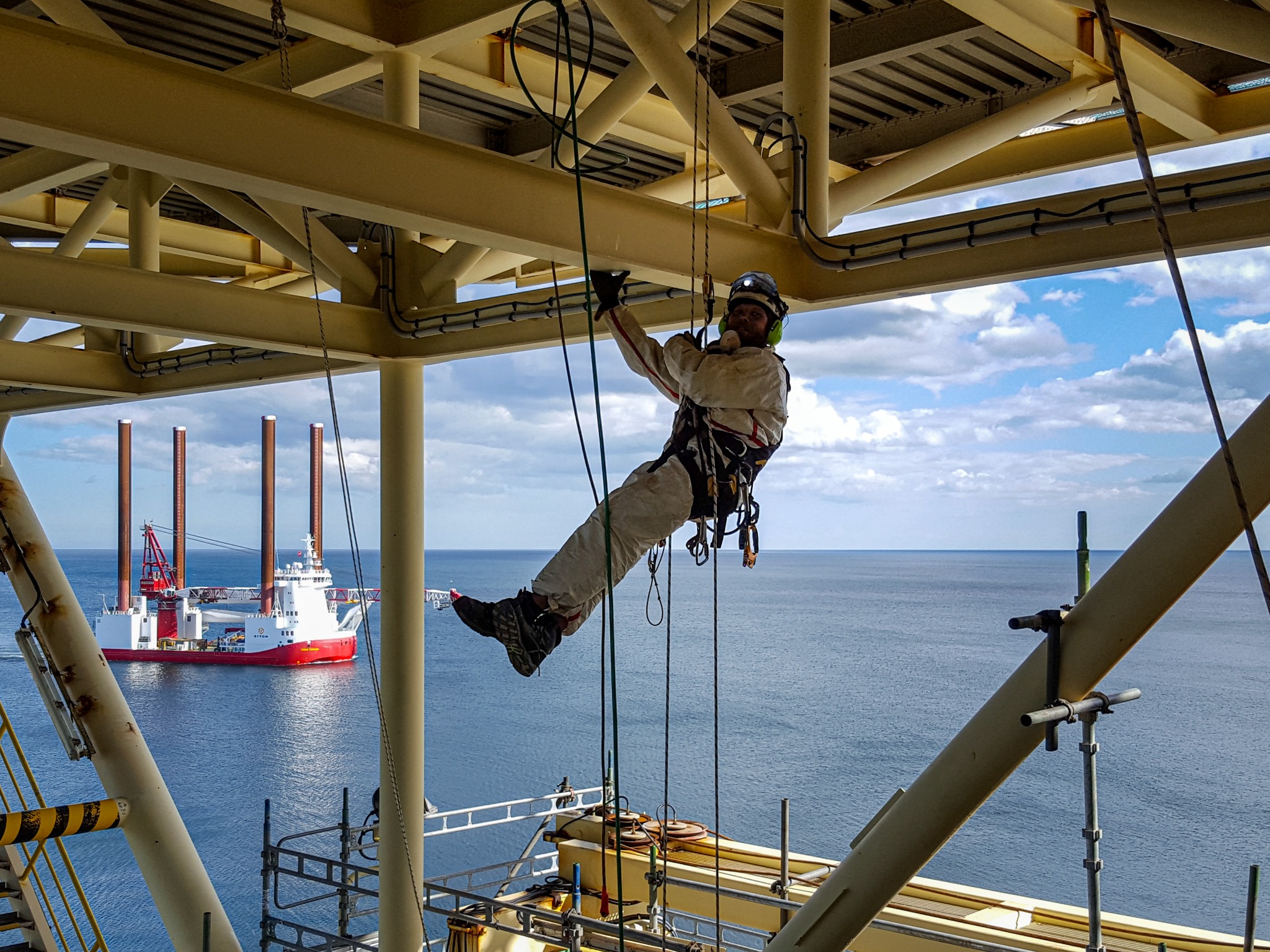 Pracownik w kombinezonie i kasku, zabezpieczony linami, wykonuje prace na wysokości na platformie wiertniczej, w tle widoczny statek i morze.