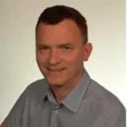 Portret uśmiechniętego mężczyzny w błękitnej koszuli na jasnym tle. Widoczne zmarszczki mimiczne i krótko ostrzyżone włosy. Dobre oświetlenie, neutralna ekspresja.