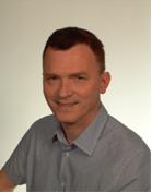 Portret uśmiechniętego mężczyzny w błękitnej koszuli na jasnym tle. Widoczne zmarszczki mimiczne i krótko ostrzyżone włosy. Dobre oświetlenie, neutralna ekspresja.