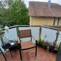 Szklana balustrada balkonu z chromowanymi poręczami, brązowymi płytkami i drewnianymi krzesłami na tle zieleni i budynku z czerwoną dachówką.