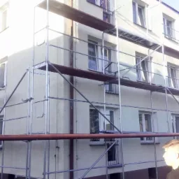 Budynek wielorodzinny w trakcie prac na elewacji, otoczony rusztowaniem, z widocznym fragmentem rury ciepłowniczej i sylwetką mężczyzny w prawym dolnym rogu.