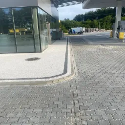 Szara, betonowa kostka brukowa ułożona na stacji benzynowej, widoczny fragment budynku stacji z przeszkloną kawiarnią i dystrybutory paliw w tle.