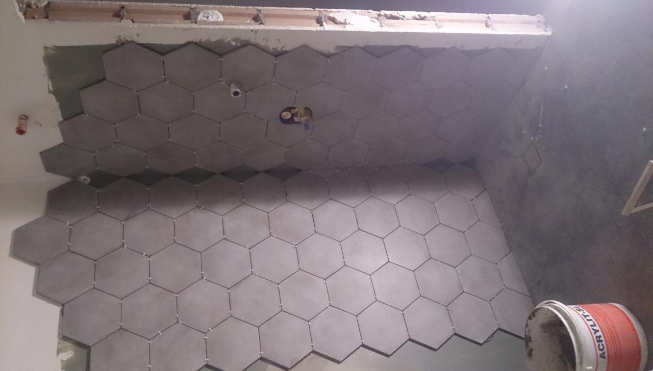 Układanie szarych, heksagonalnych płytek na ścianie w łazience. Widoczne krzyżyki dystansowe i puszka z klejem. Etap prac wykończeniowych w trakcie remontu.