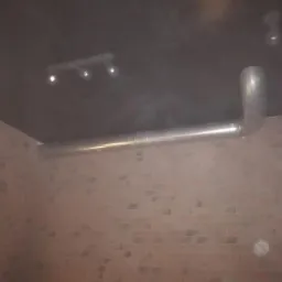 Metalowa rura wentylacyjna poprowadzona wzdłuż ceglanej ściany pod ciemnym sufitem z reflektorami punktowymi.