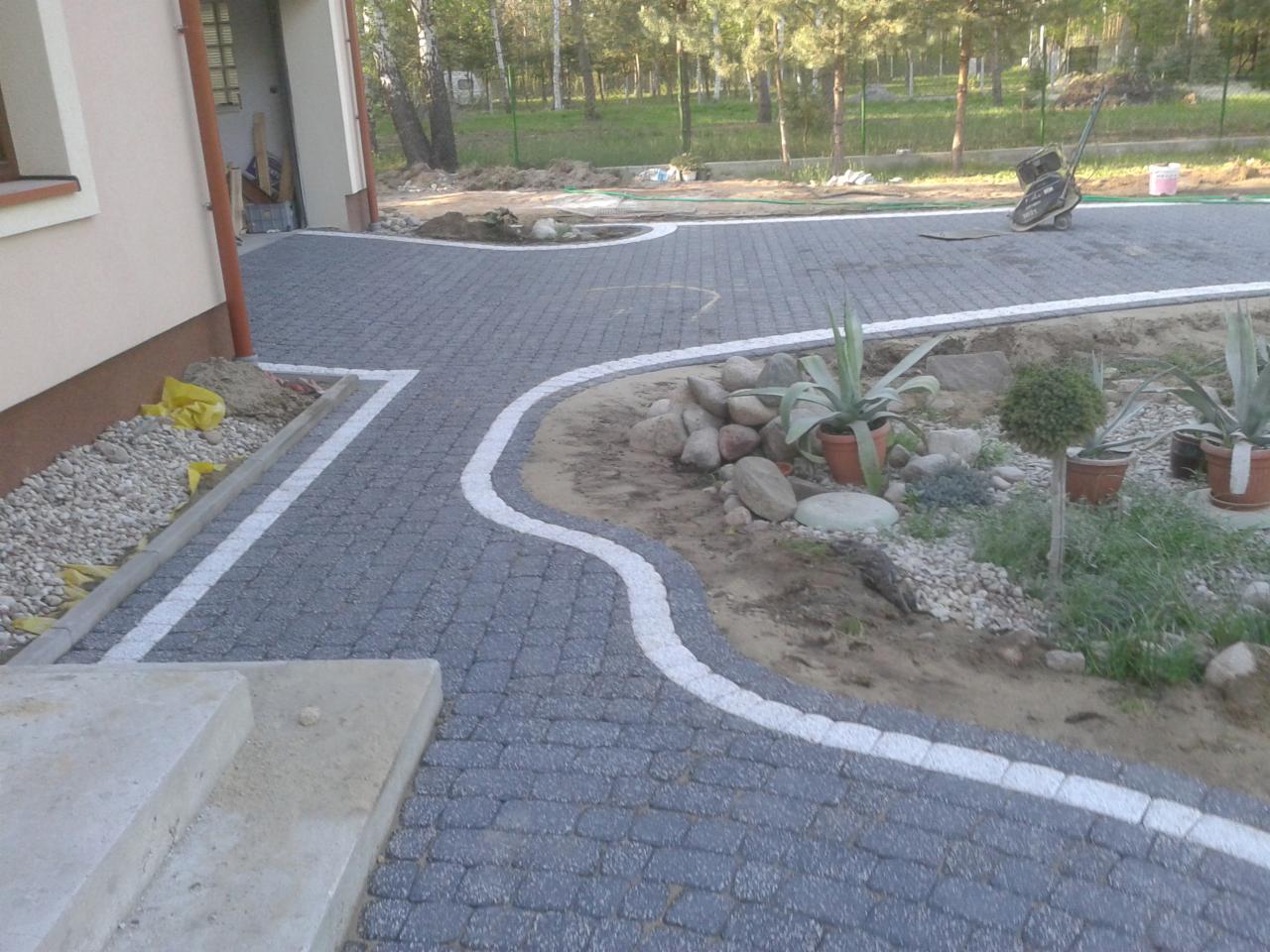 Szara kostka brukowa z białym obramowaniem prowadząca od domu do ogrodu. W tle widoczna maszyna do ubijania. Rośliny w donicach dodają uroku.
