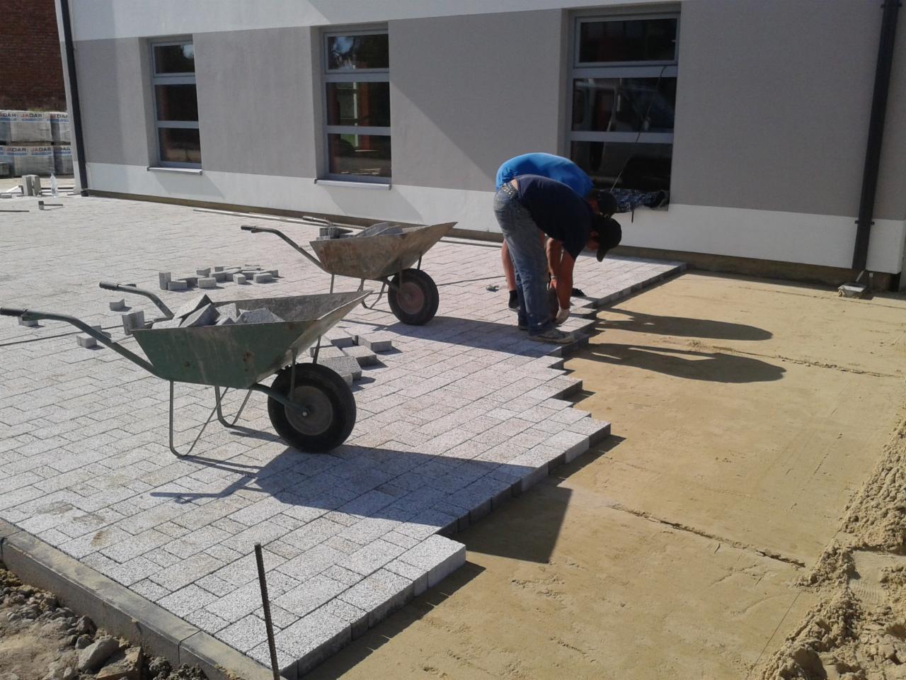 Dwóch brukarzy układa szarą kostkę na podwórku obok budynku. Dwie taczki wypełnione kostką stoją obok. Częściowo ułożona nawierzchnia z kostki.