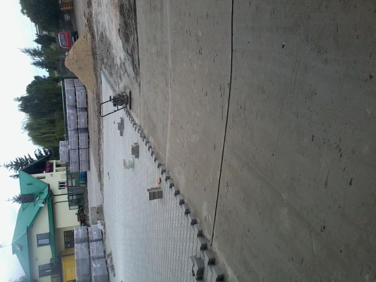 Układanie szarej kostki brukowej na posesji, widoczna ubijarka, palety z kostką, dom z zielonym dachem w tle.