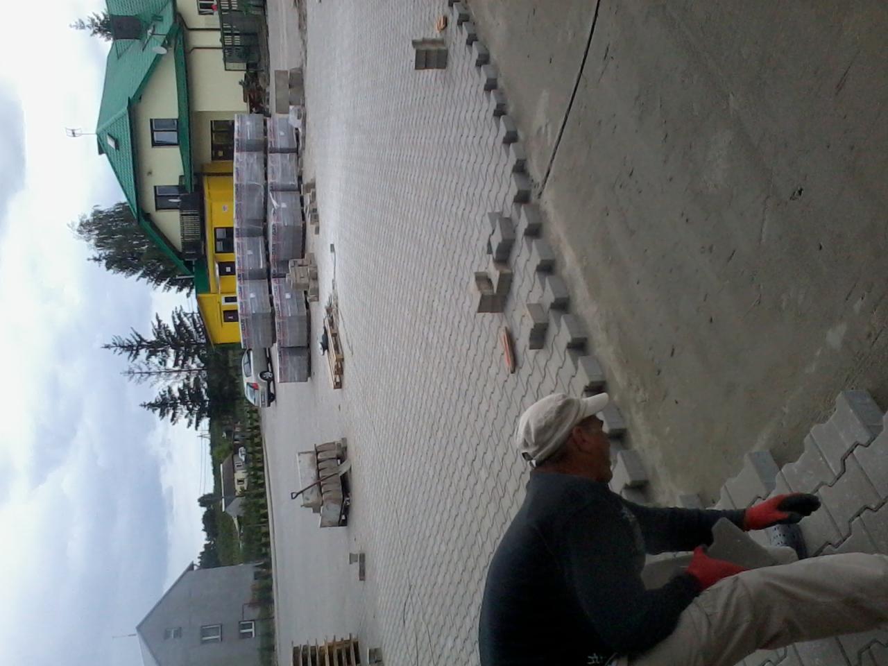 Pracownik w czerwonych rękawicach układa zygzakowatą kostkę brukową na przygotowanej powierzchni, z widocznymi paletami kostki w tle i budynkami w oddali.