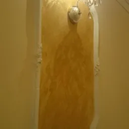 Dekoracyjna ściana z efektem złota, obramowana białym ornamentem i oświetlona kryształowym kinkietem, tworzy elegancki akcent w jasnym wnętrzu.