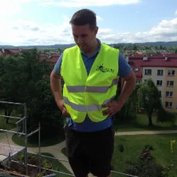 Pracownik w kamizelce Global Sun na dachu budynku z rusztowaniem, w tle zabudowa miejska i zieleń. Kadr z góry, widok na instalację.