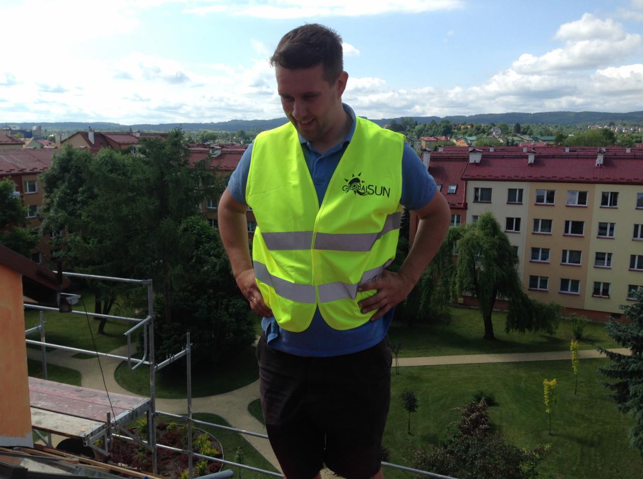 Pracownik w kamizelce Global Sun na dachu budynku z rusztowaniem, w tle zabudowa miejska i zieleń. Kadr z góry, widok na instalację.