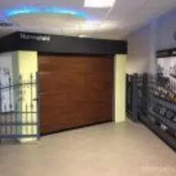 Prezentacja segmentowej bramy garażowej w kolorze drewna, umieszczonej w salonie ekspozycyjnym z widocznymi elementami ogrodzeń po bokach.