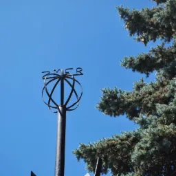 Metalowa konstrukcja w kształcie kuli z napisem 'ZMER' na szczycie słupa, obok rozłożystego drzewa iglastego na tle błękitnego nieba.