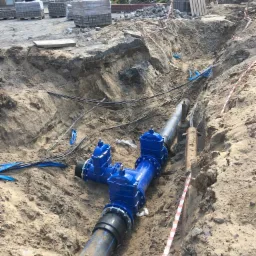 Wykop w ziemi z widocznym, częściowo zakopanym, niebieskim przyłączem wodociągowym z zaworami, w tle kostka brukowa i budynki handlowe.