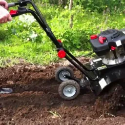 Czarna glebogryzarka spalinowa w trakcie pracy, operator w jeansach i koszuli w kratę, świeżo przekopana ziemia w ogrodzie.