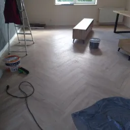 Nowo ułożona podłoga z paneli w jodełkę, widoczne akcesoria malarskie, drabina i meble w jasnym pokoju.