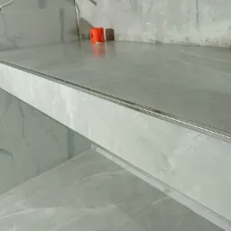 Półka wykończona szarymi płytkami imitującymi marmur, z metalową listwą na krawędzi, widoczne gniazdko elektryczne i puszki instalacyjne na surowej ścianie.