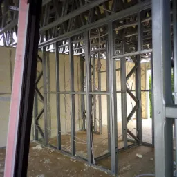 Wnętrze budynku w trakcie budowy, widoczna metalowa konstrukcja szkieletowa ścian i stropu, częściowo wypełniona jasną pianą izolacyjną, z widocznymi przewodami elektrycznymi.