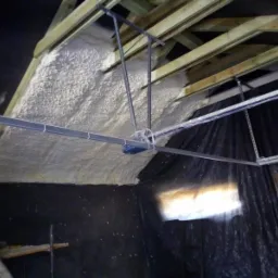 Poddasze w trakcie izolacji natryskową pianką, widoczne drewniane krokwie, folia ochronna na ścianach i metalowa konstrukcja pod sufitem.