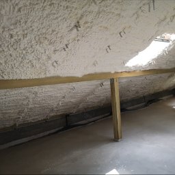 EDWARD FEŃSKI ~EDMAR~ - Poddasze ocieplone pianką PUR, widoczne drewniane belki konstrukcyjne i metalowe mocowania. Biała, nierówna powierzchnia pianki pokrywa skośny dach.