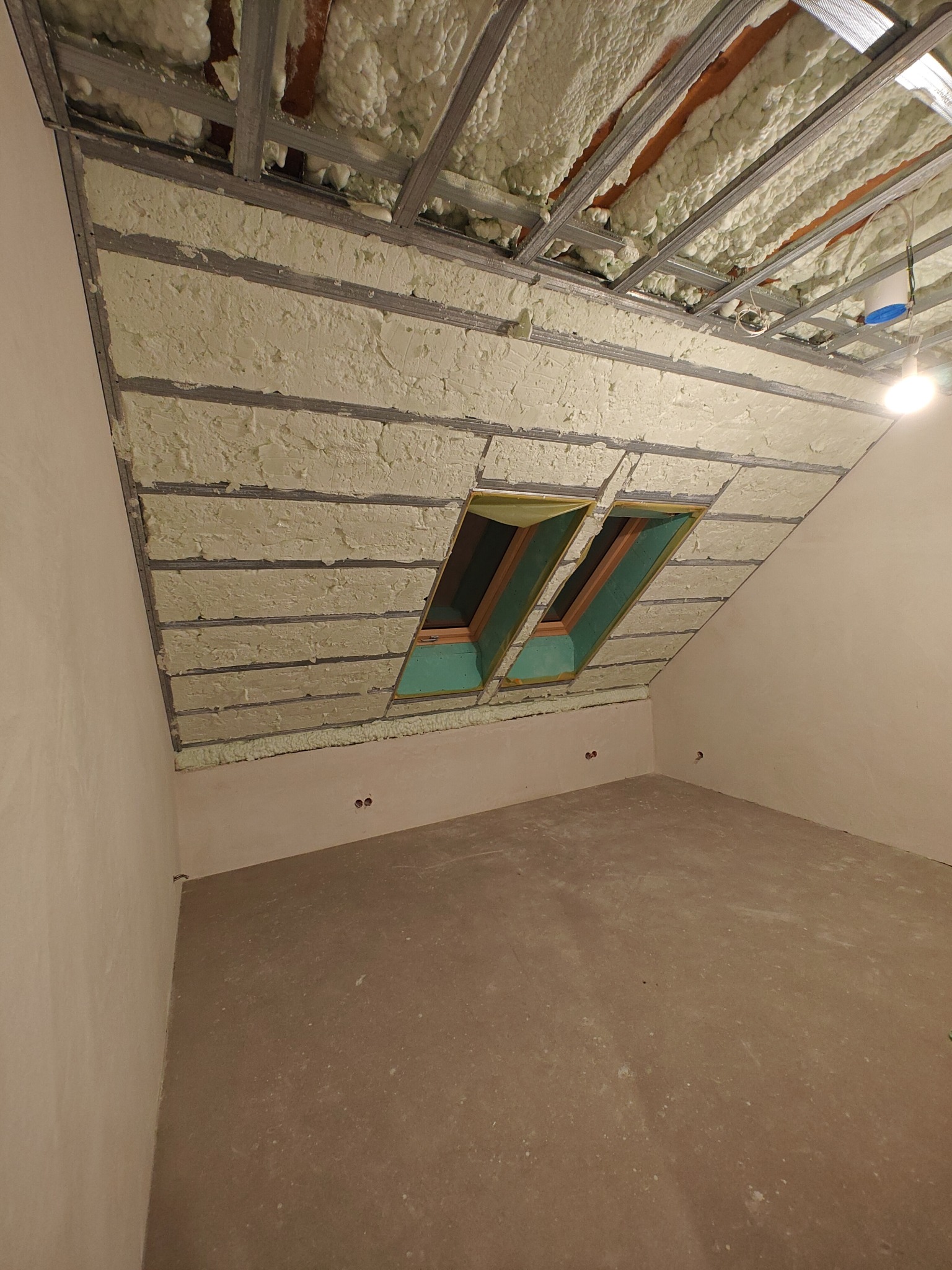 Poddasze po ociepleniu pianką PUR, widoczne metalowe profile i dwa okna dachowe. Ściany przygotowane do wykończenia, surowa podłoga. Oświetlenie halogenowe.