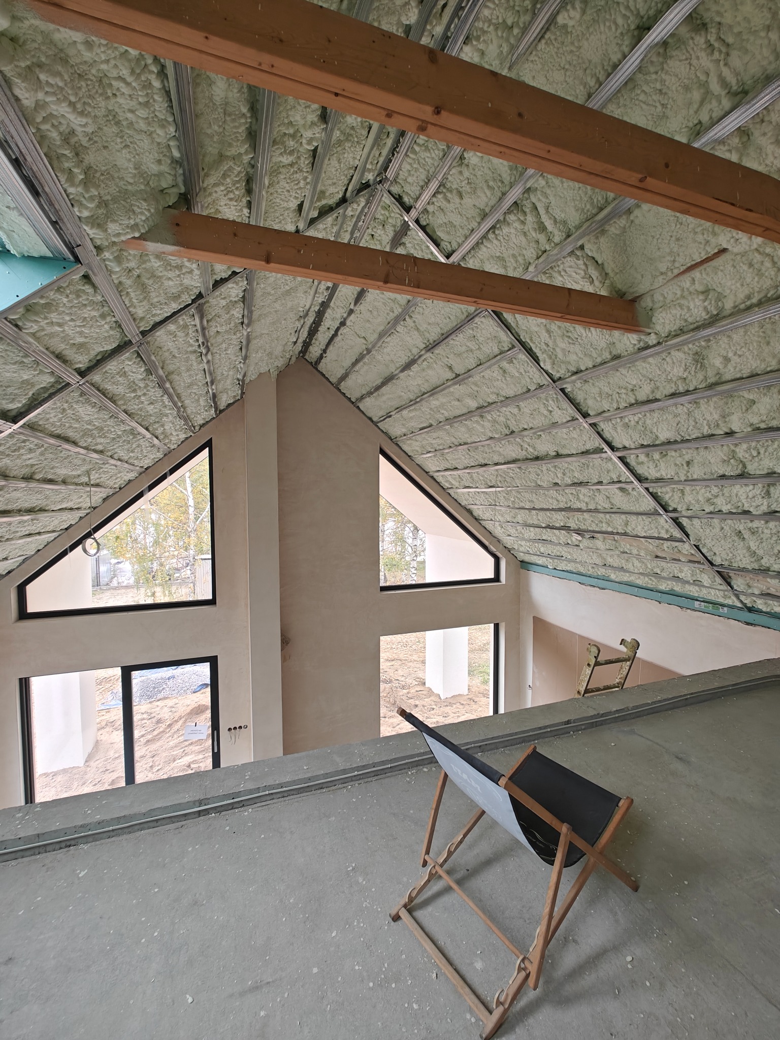 Poddasze domu z oknami szczytowymi, ocieplone pianką PUR, widoczne drewniane belki i metalowa konstrukcja sufitu. Na pierwszym planie leżak.