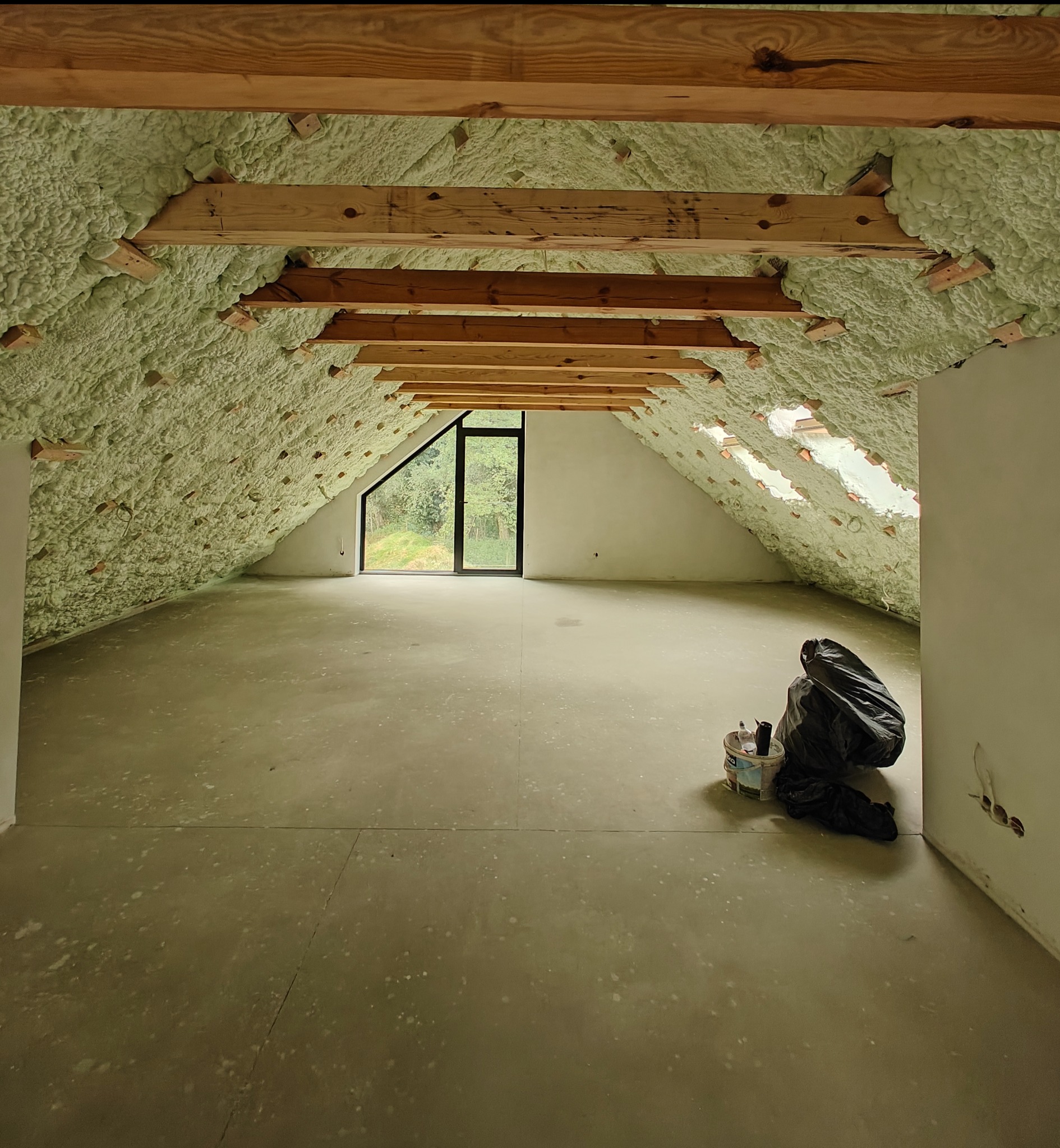 Poddasze po ociepleniu pianką PUR, widoczne drewniane belki i okno. Na podłodze wiadro i worek. Ściany białe, surowe. Ocieplenie wykonane starannie, pokrywa całą powierzchnię.