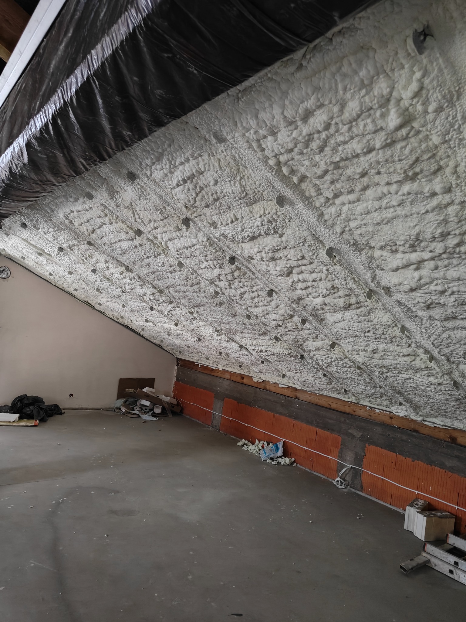 Poddasze po ociepleniu pianką PUR: widoczne białe, nieregularne struktury pianki na skośnym dachu, czarna folia w górnej części, ceglana ściana boczna, surowy stan wykończenia.