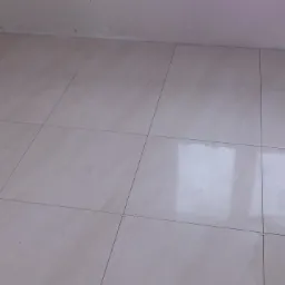 Jasne, beżowe płytki podłogowe ułożone w regularny wzór, widoczne fugi i odbicie światła, ściany w tle w odcieniach bieli, minimalistyczne wykończenie wnętrza.