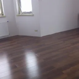 Ułożona, brązowa podłoga z paneli w jasnym pokoju z grzejnikami. Okna zabezpieczone folią. Widoczne odbicia światła na powierzchni paneli.