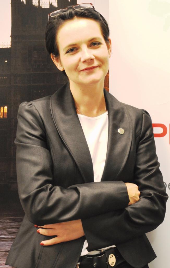 Portret kobiety w ciemnej marynarce, z rękami skrzyżowanymi na piersiach, w tle Big Ben. Okulary na głowie, delikatny uśmiech. Profesjonalny wizerunek.